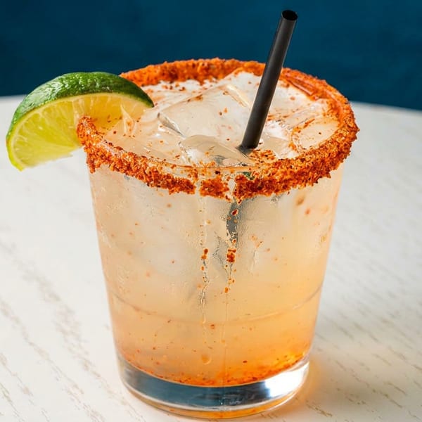 A spicy margarita with a chili-salt rim, lime wedge, and black straw on a white table.