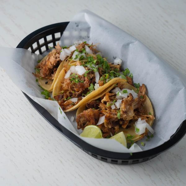 Three carnitas tacos with diced onions, cilantro, and lime wedges in a black basket.