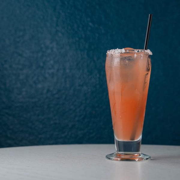 Tall glass of pink cocktail with a salted rim and black straw on a white table.