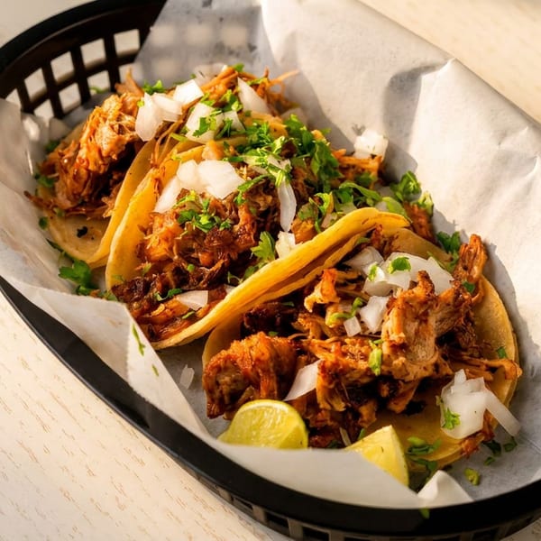 Three carnitas tacos with onions, cilantro, and lime wedges in a black basket on a wooden table.