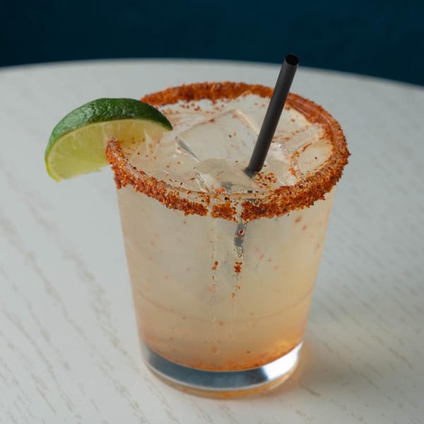A light-colored cocktail with a chili-lime rim, ice, a black straw, and a lime wedge.