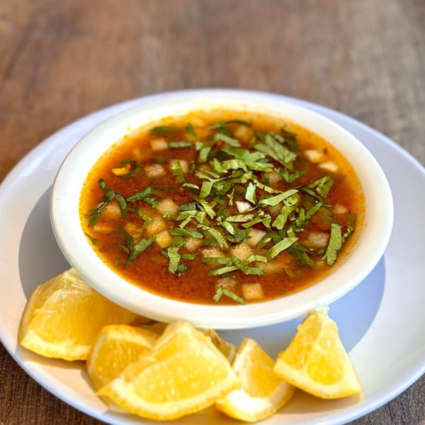 A white bowl of red soup with diced ingredients and green herbs, served with lemon wedges.