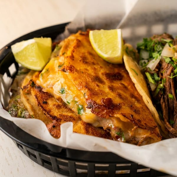 A black basket holds two birria tacos with lime wedges and two street tacos with cilantro and onions.