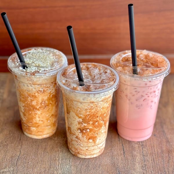 Three iced drinks, two brown and one pink, with black straws on a wooden surface.